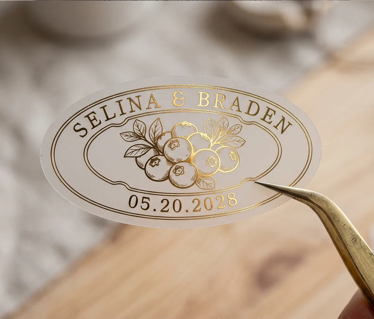 Oval personalized gold foil print matte wedding favour label held up by gold tweezer.