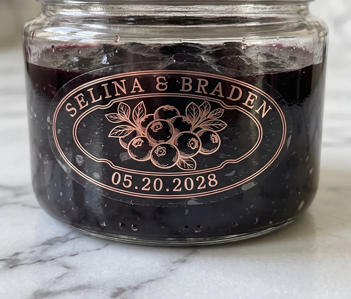 Oval personalized rose gold foil print clear wedding favour label on a blueberry jam jar.