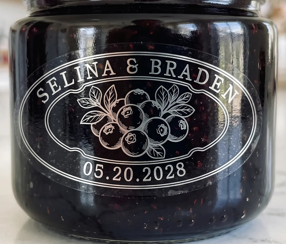 Oval personalized silver foil print clear wedding favour label on a blueberry jam jar.