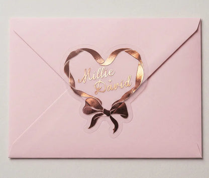 Ribbon in heart shape wedding labels with couples names printed in rose gold foil on transparent frosted vinyl sealing an envelope