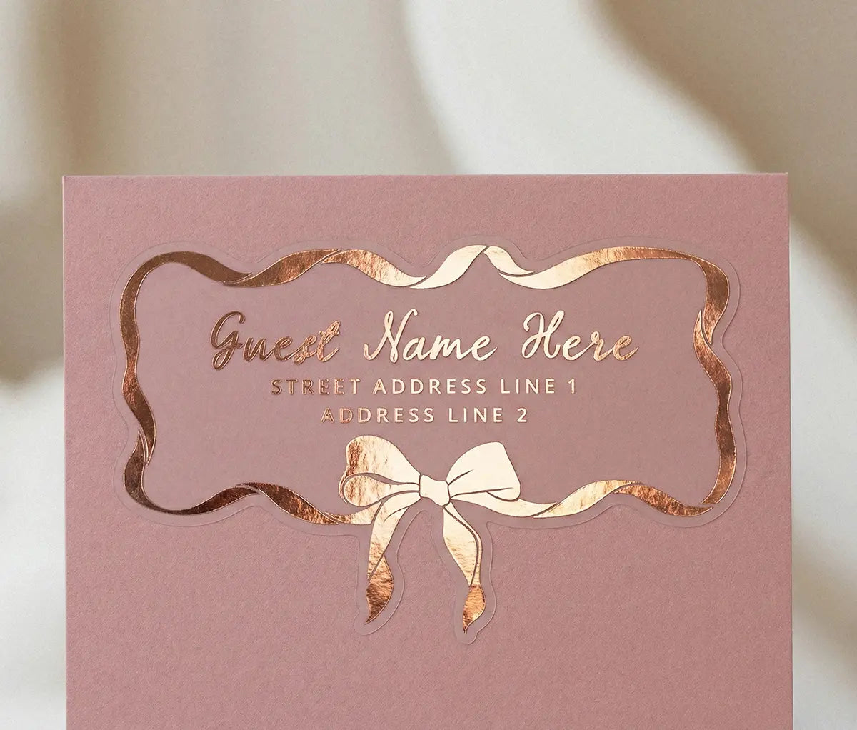 Guest address label printed in rose gold foil applied to dusty-pink color envelope with custom die-cut coquette bow and ribbon shape.
