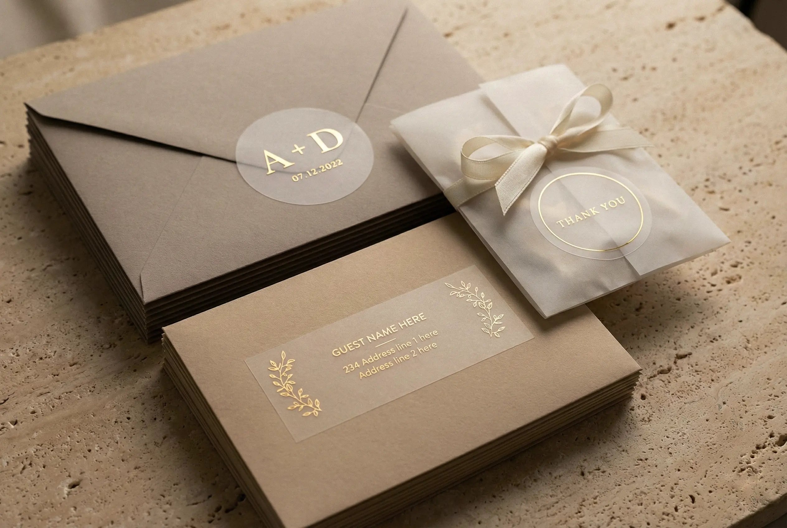 A photograph highlights three gold-foiled clear stickers used as seals and labels on beige wedding stationery. On the top left, a round sticker with gold foil initials "A + D" and "07.12.2022" seals a stack of envelopes. On the bottom left, a rectangular sticker with gold foil text "GUEST NAME HERE," address lines, and leaf designs labels another stack of envelopes. On the right, a round sticker with "THANK YOU" in gold foil is affixed to a small vellum packet tied with a white ribbon.