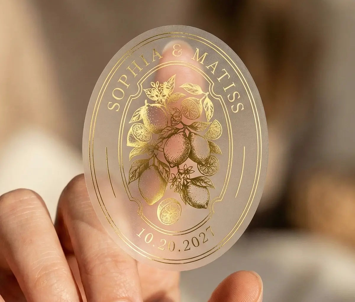 Transparent matte oval sticker with lemon graphic and custom names and date stuck in the index finger.