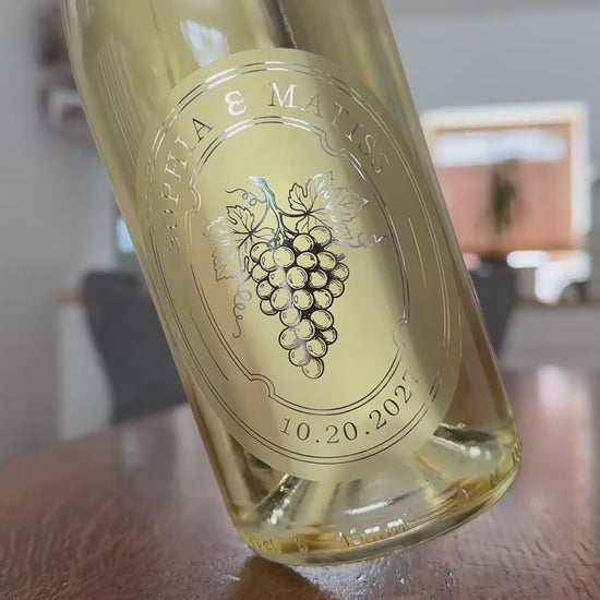Personalized frosted wedding favour sticker applied to a white wine bottle. Bottle is being rotted slightly to show the shine of the silver foil print.