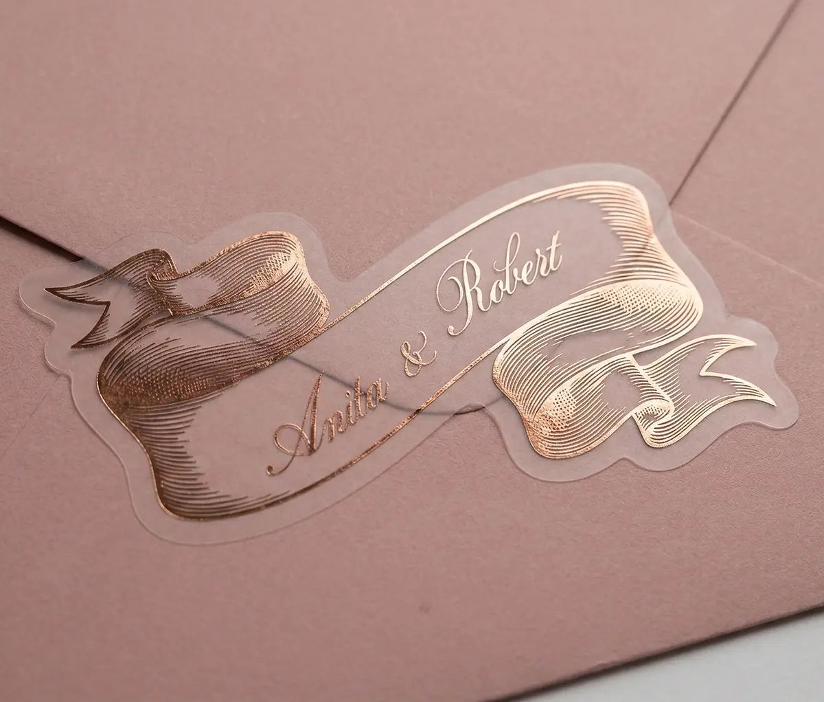 Wedding sticker cut in vintage style ribbon shape with personalized names and rose gold foil print on a pink envelope.