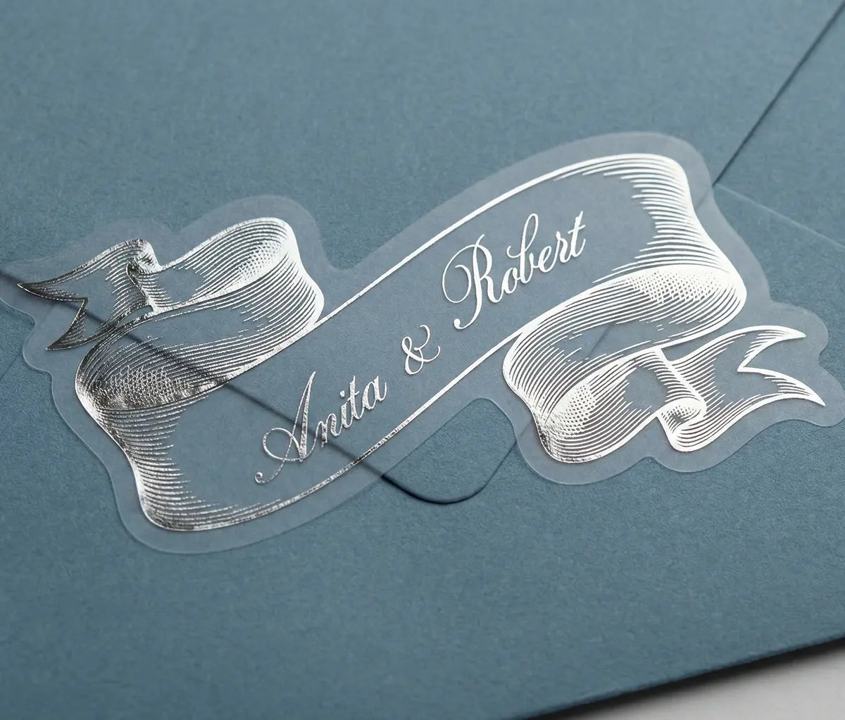 Vintage style ribbon shape wedding stic.ker with custom names printed in silver foil applied to blue envelope