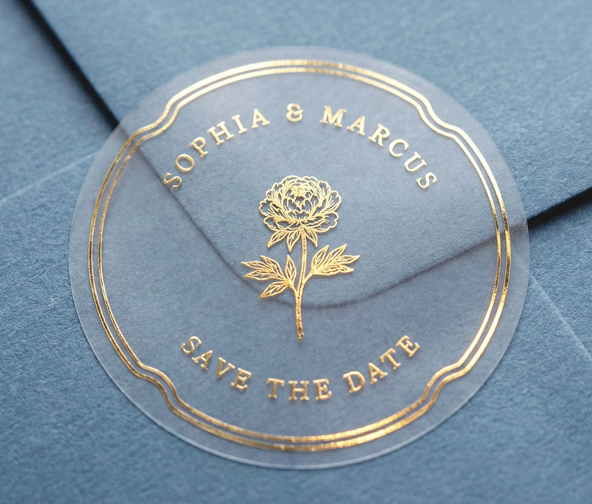 Extreme close-up of a transparent Save the Date sticker with peony graphic and personalized names printed in gold foil sealing slate blue envelope.