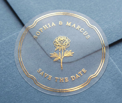 Extreme close-up of a transparent Save the Date sticker with peony graphic and personalized names printed in gold foil sealing slate blue envelope.