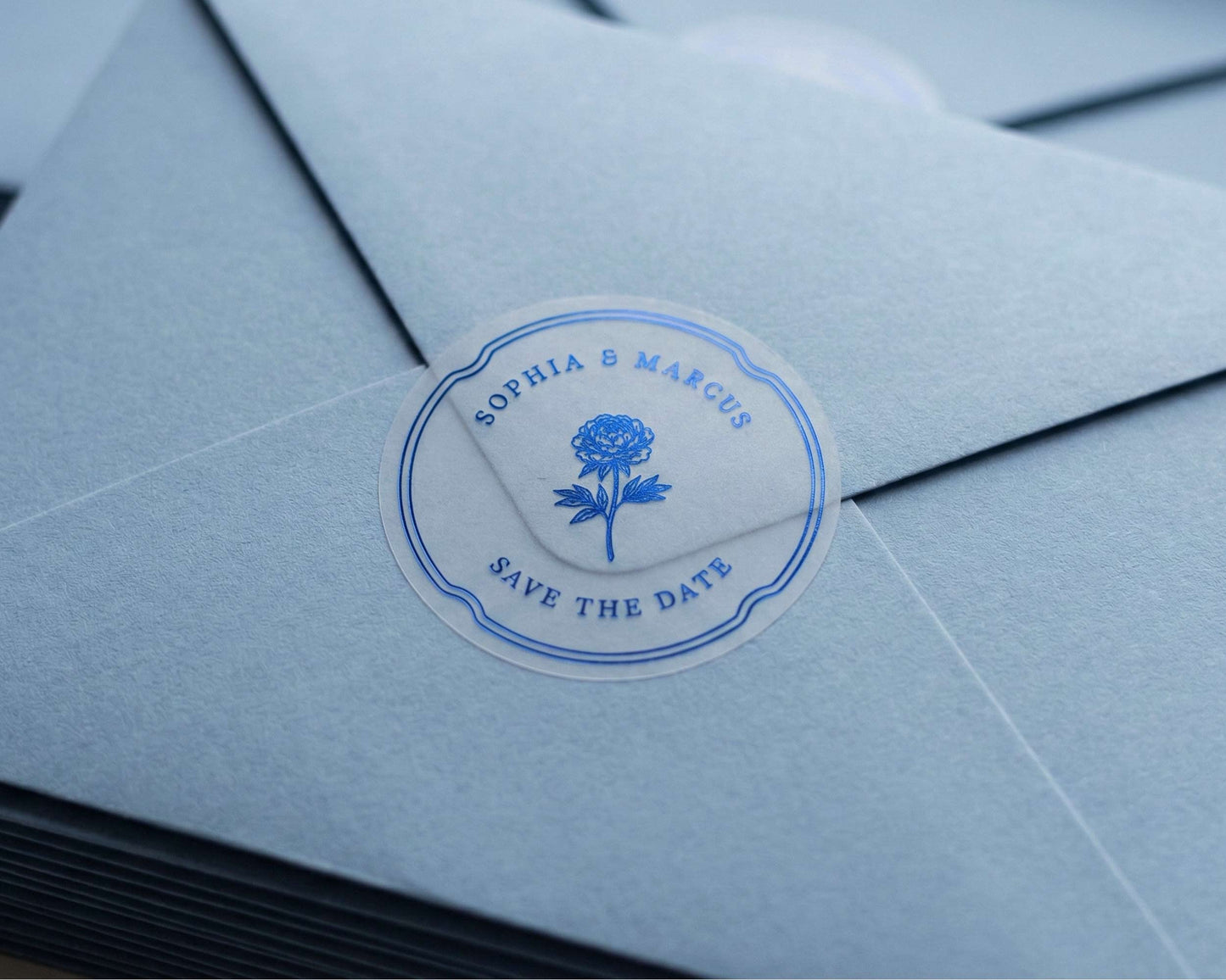 Clear Save the Date sticker with flower graphic and personalized names printed in royal blue foil sealing light blue envelope.