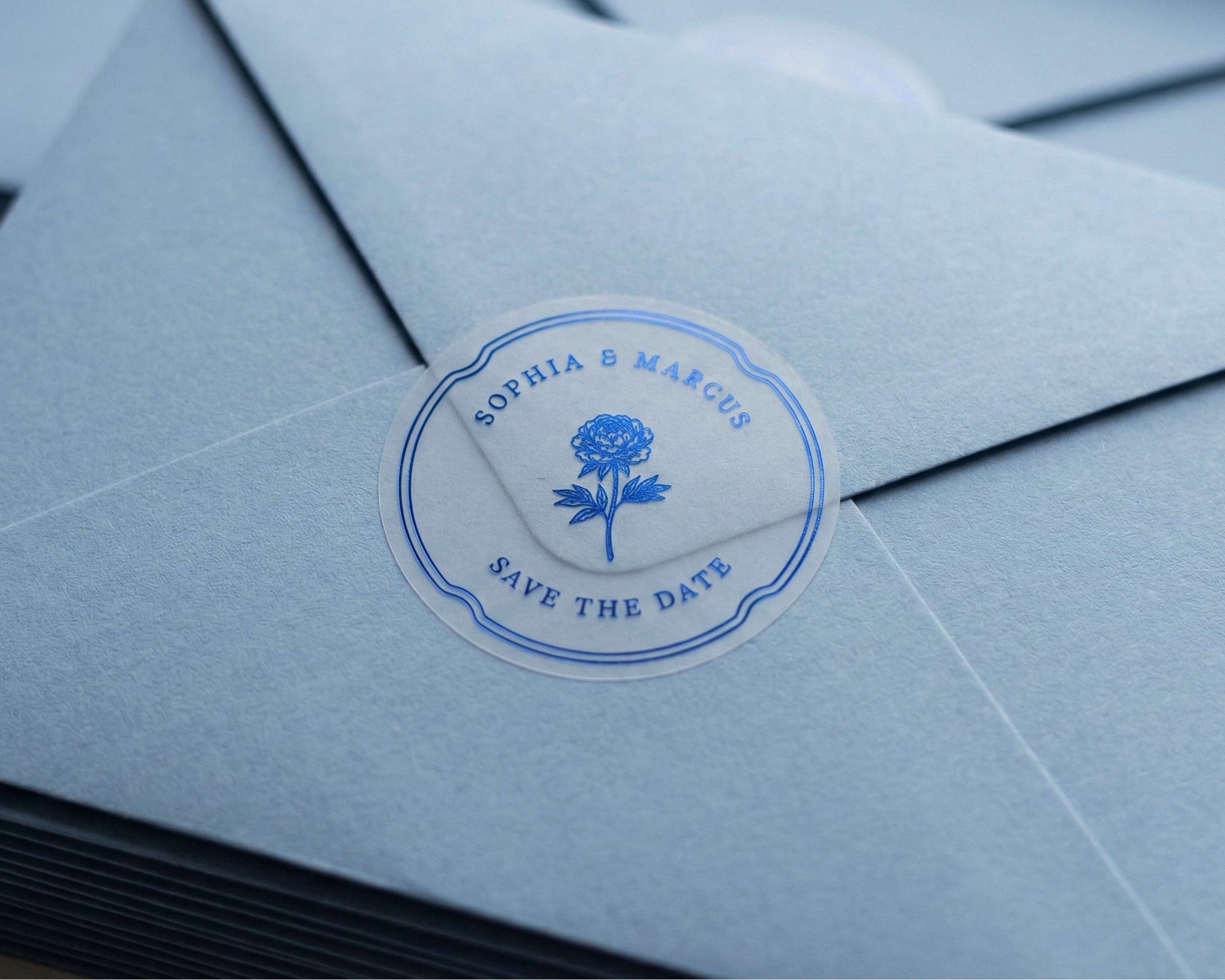 Clear Save the Date sticker with flower graphic and personalized names printed in royal blue foil sealing light blue envelope.