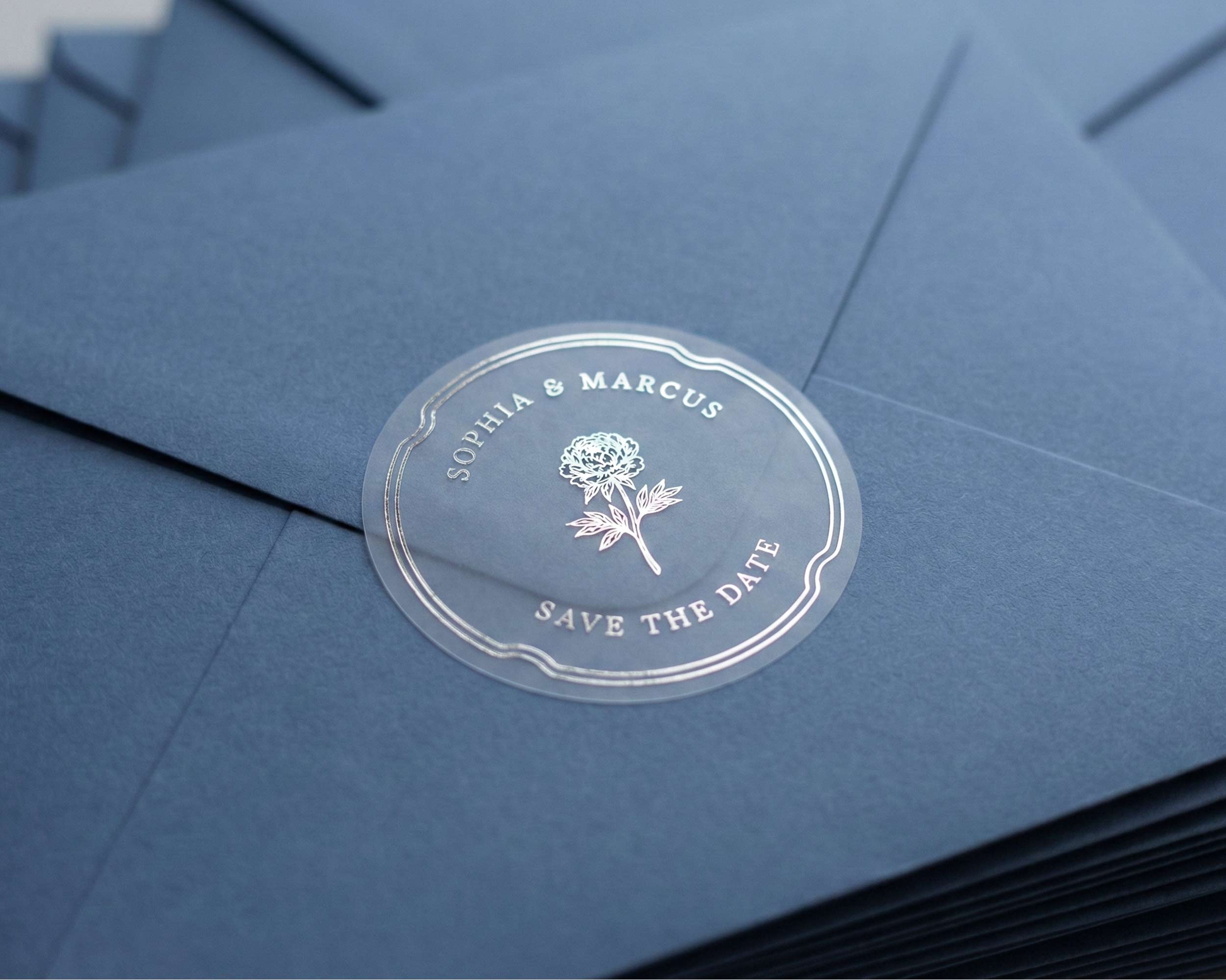 Clear Save the Date sticker with flower graphic and personalized names printed in silver foil sealing navy blue envelope.
