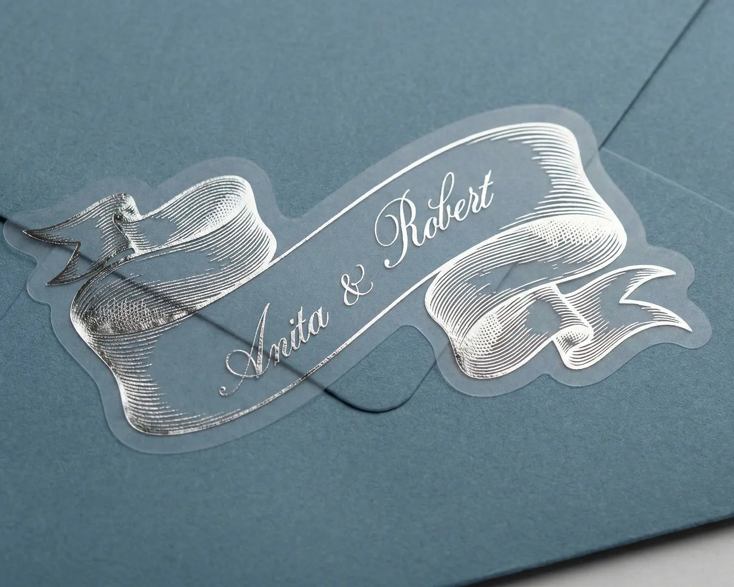 Decorative ribbon wedding envelope sticker with custom text 'Anita & Robert' on a blue envelope.